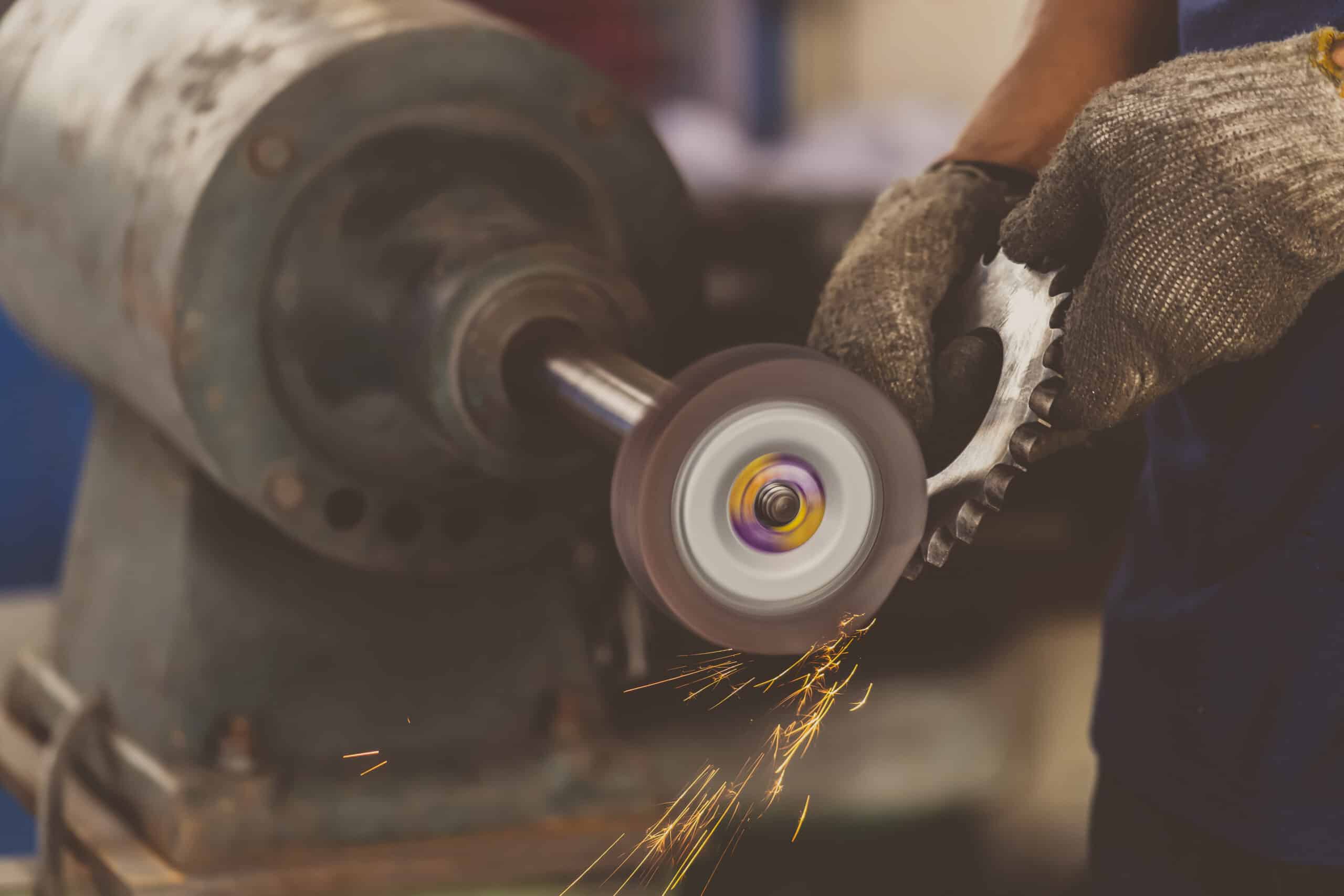 Mit Handschuhen gehaltenes Metallzahnrad wird gegen ein rotierendes Schleifrad geschliffen, wobei Funken sprühen und ein unscharfer Werkstattmotor im Hintergrund zu sehen ist.