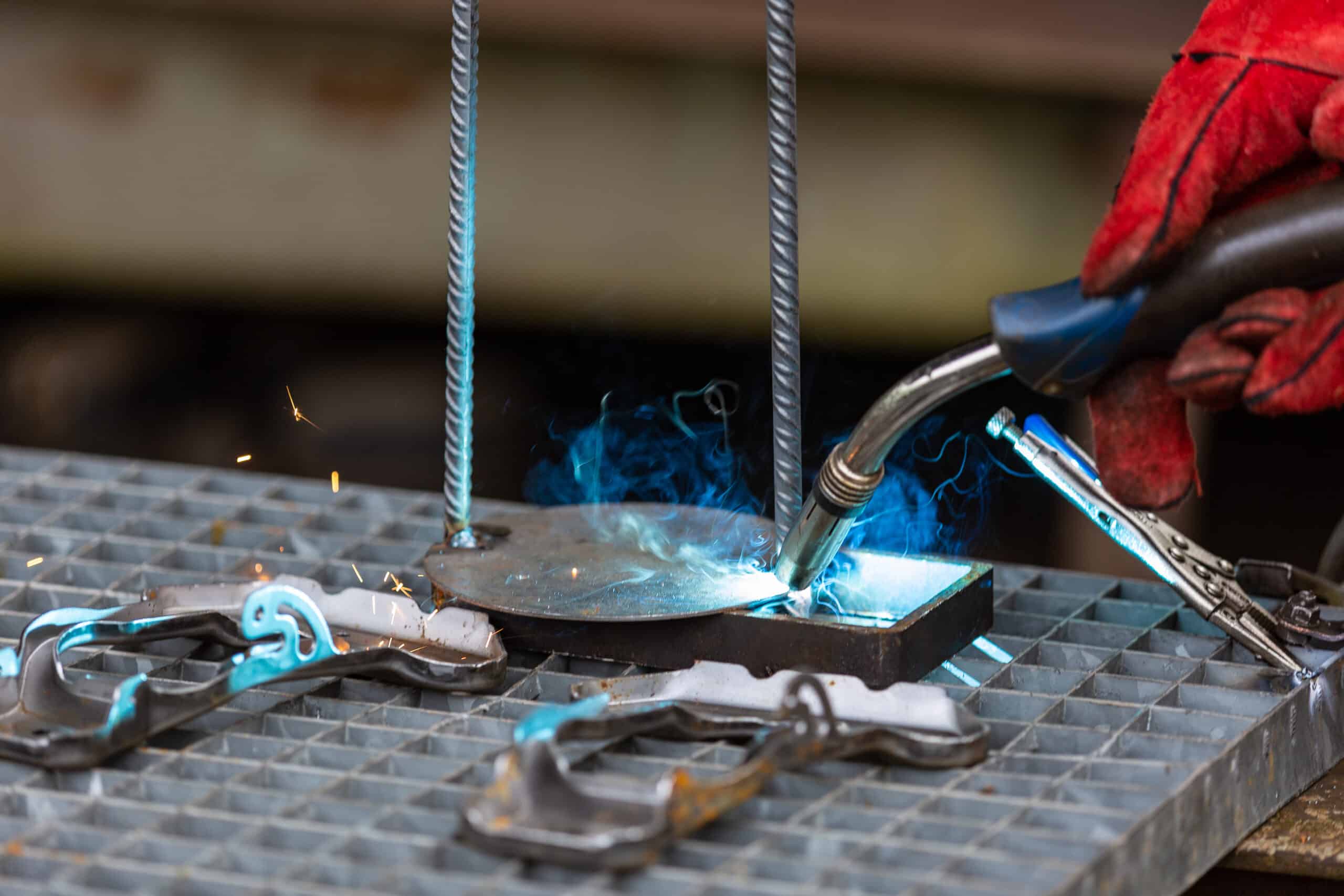 Hand mit rotem Schutzhandschuh führt einen Schweißbrenner, der eine runde Metallplatte auf einem rechteckigen Auflageblech auf einem Gittertisch verschweißt; blaue Funken und Rauch sowie Metallklammern und zwei vertikale Bewehrungsstäbe sind sichtbar.