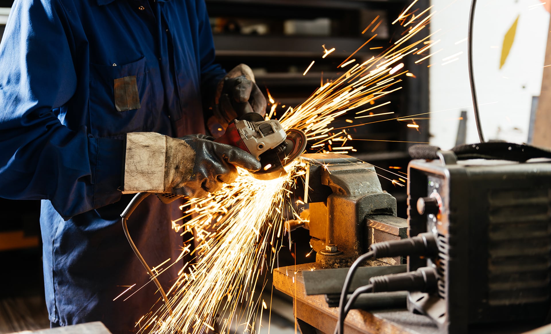 Arbeiter in blauer Schutzkleidung und dicken Handschuhen schleift ein Metallstück mit einem Winkelschleifer im Schraubstock, wobei helle Funken in einer Werkstatt sprühen.
