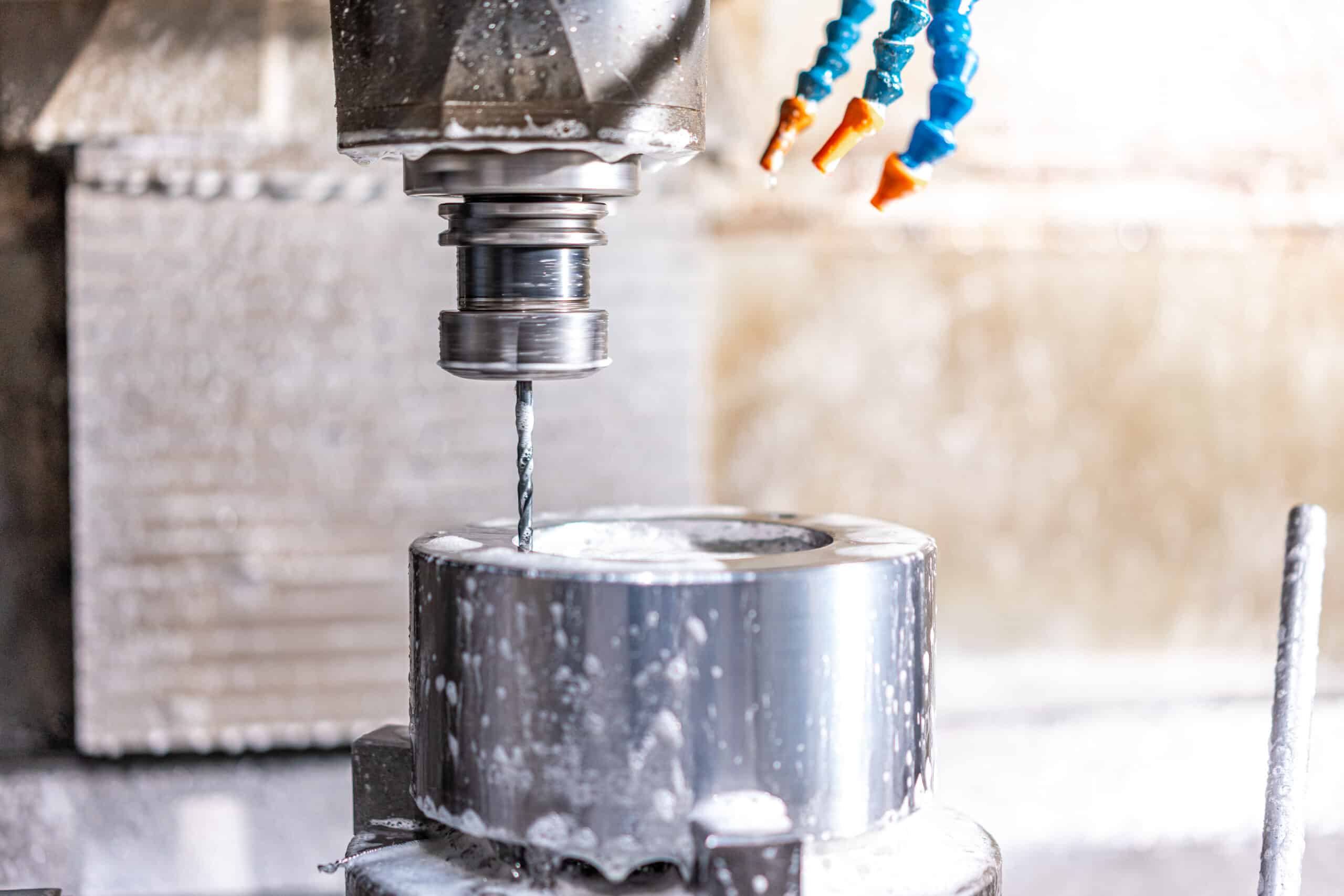 Nahaufnahme einer CNC‑Bohrspindel mit feinem Bohrer, der ein Loch in einen zylindrischen Metallblock fräst; sichtbar sind Kühlflüssigkeitsspritzer und blaue-orange flexible Kühlrohre.