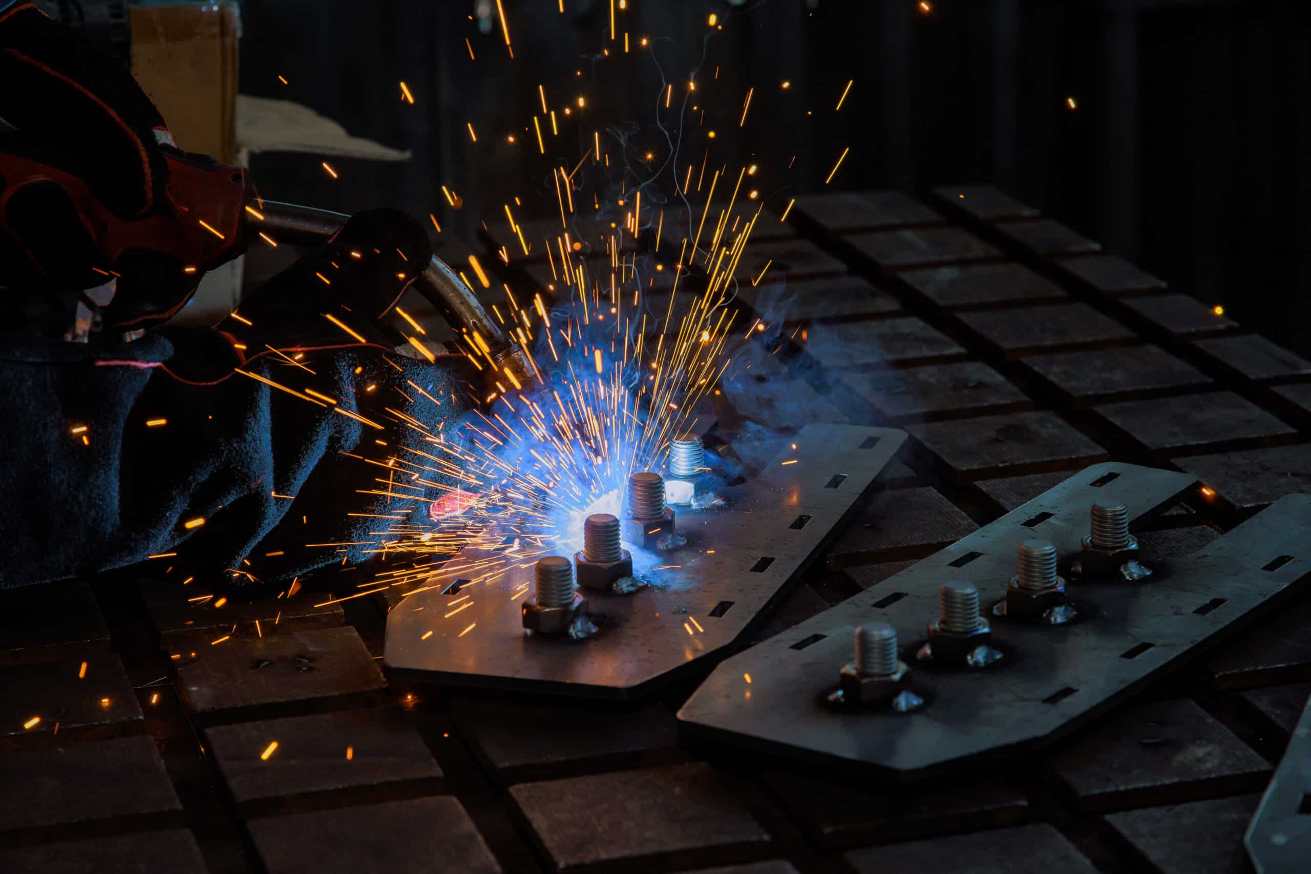 Behandschuhte Hand hält einen Schweißbrenner über Metallplatten mit aufgeschraubten Bolzen; blaue Flamme und goldene Funken sprühen über eine dunkle Werkbank.