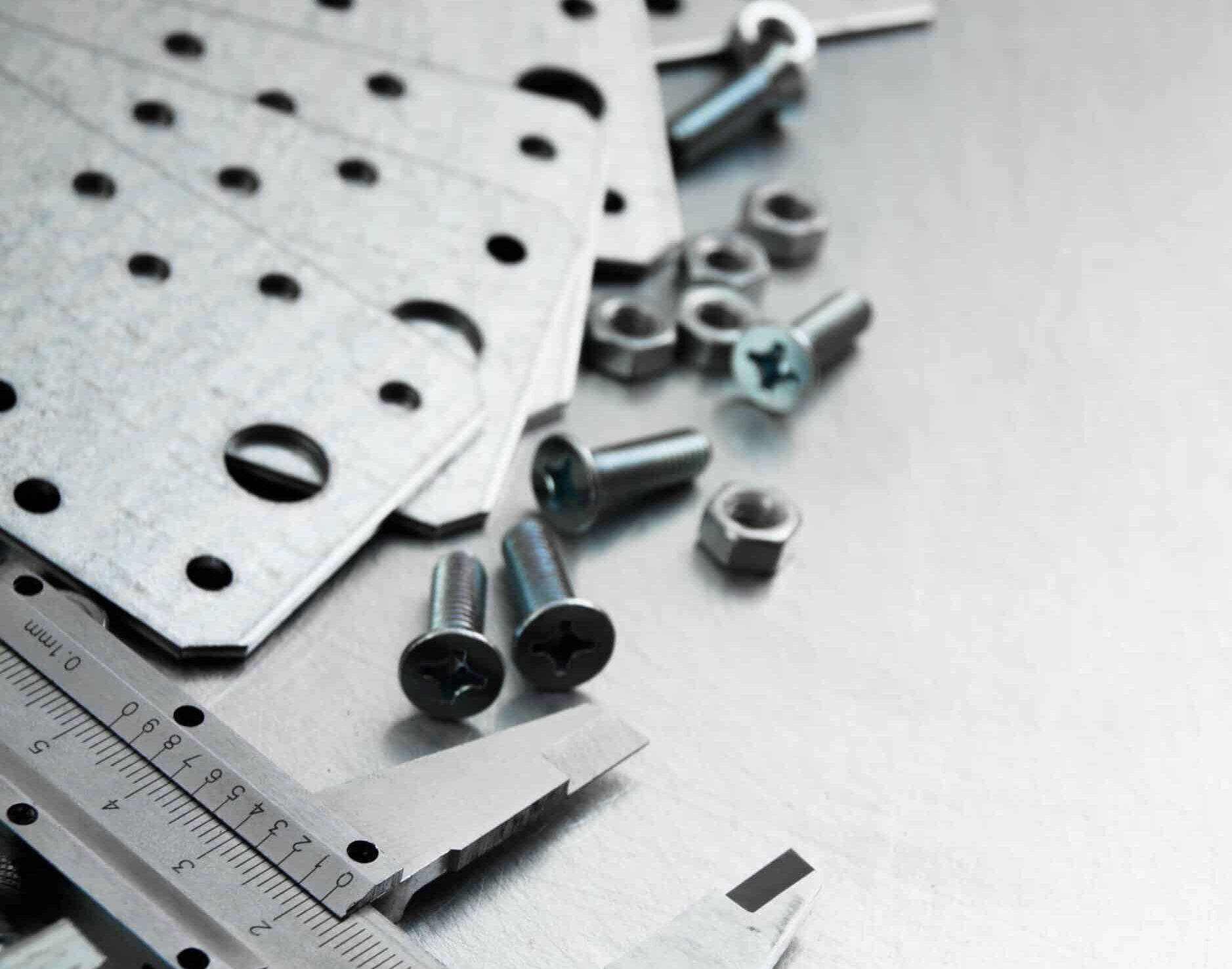 Verschiedene verzinkte Metallverbinderplatten mit runden Bohrungen, mehrere Kreuzschlitzschrauben und Muttern sowie ein teilweiser Messschieber liegen verstreut auf einer gebürsteten Metalloberfläche.