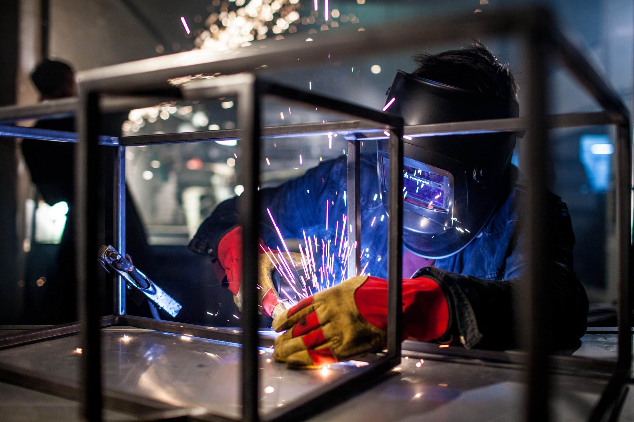 Person mit Schutzhelm und roten Lederhandschuhen schweißt an einem rechteckigen Metallrahmen, während helle Funken und blaues Licht in einer Werkstatt aufleuchten.