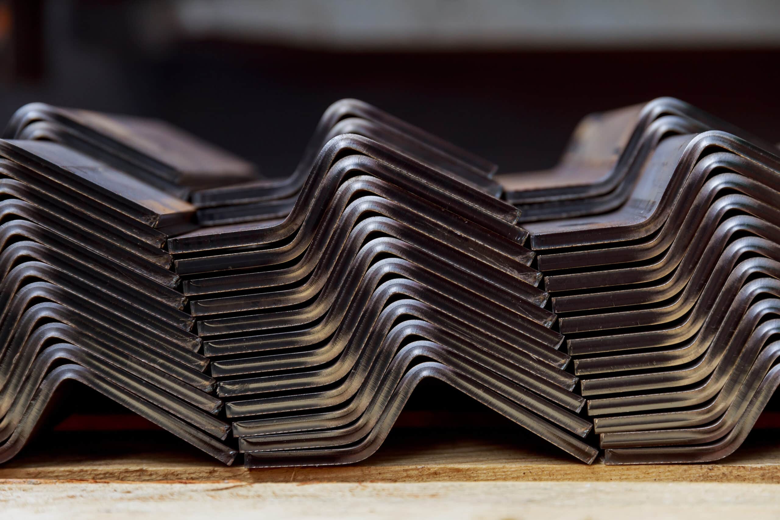 Mehrere dunkel glänzende, z‑förmig gebogene Metallbleche dicht übereinander gestapelt auf einer Holzunterlage.